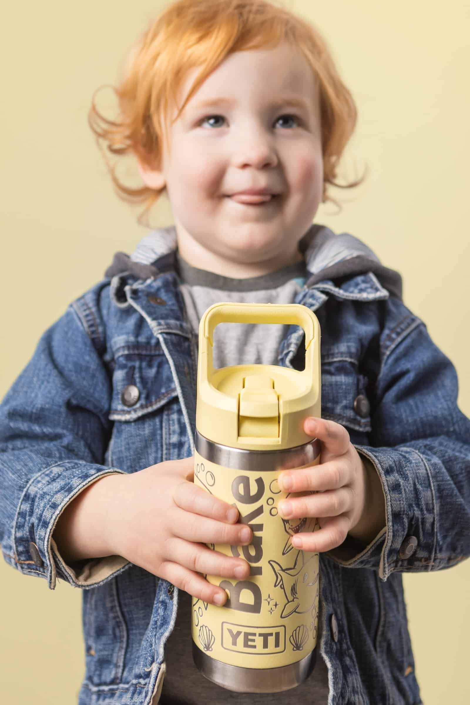 Young child with red hair and blue eyes smiling, holding a yellow YETI water bottle, dressed casually.