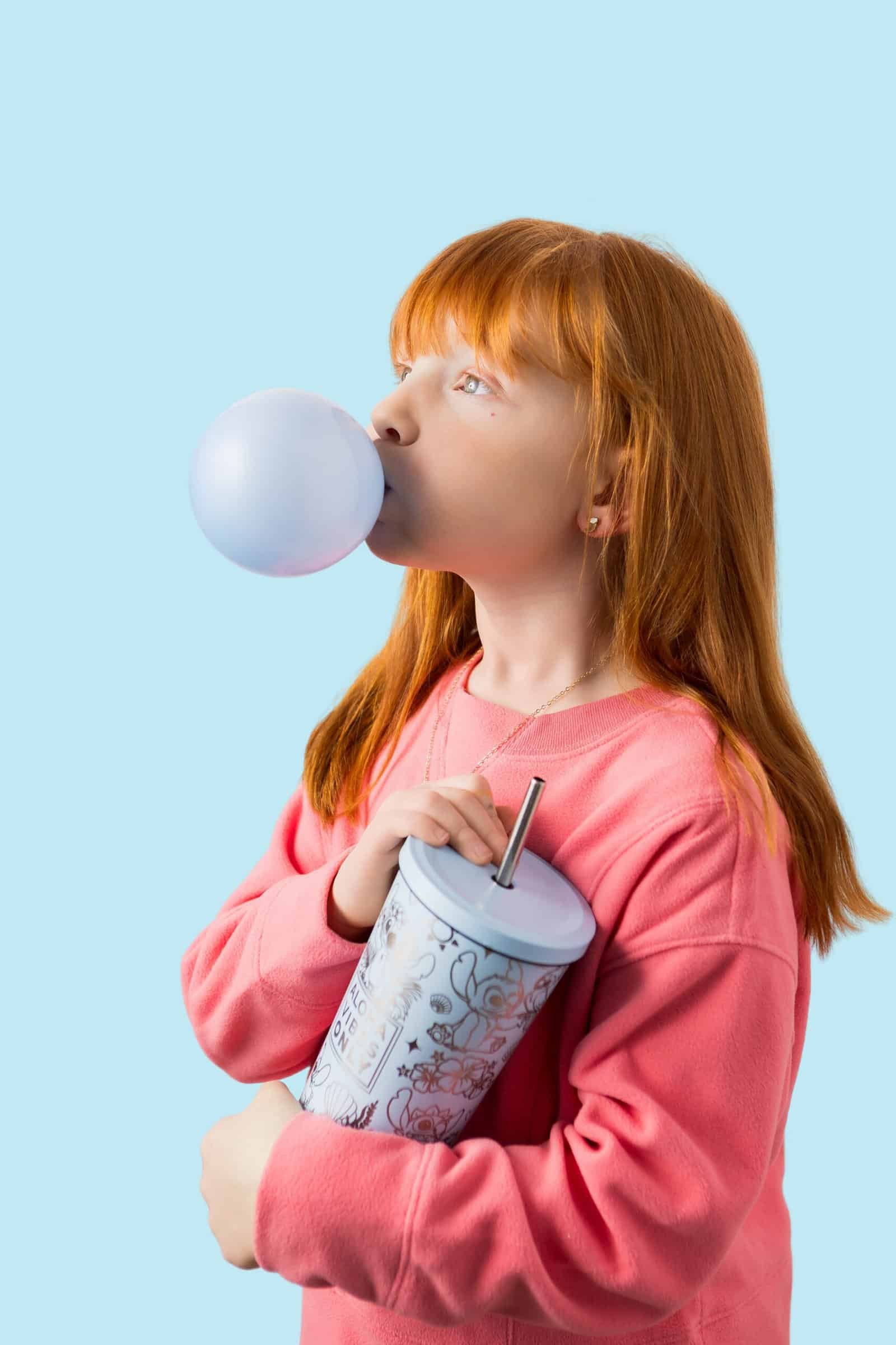 Young girl blowing bubblegum with a drink cup against a light blue background.